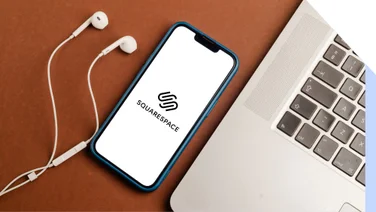 brown tables with white earphones, phone with squarespace's logo, and a laptop on it