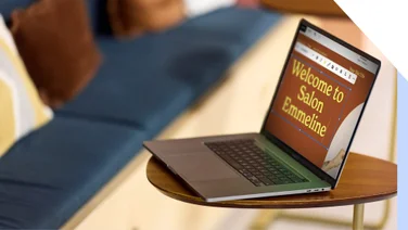 Open laptop on a wooden table displaying 'Welcome to Salon Emmeline' Squarespace template on screen with a blurred sofa in the background.