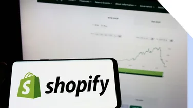 A phone with the shopify logo on it in front of a laptop screen with a graph