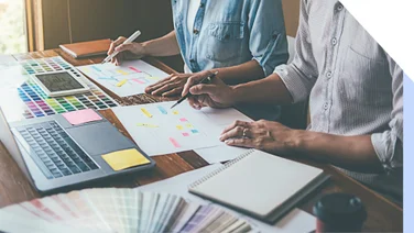 Two people sit at a desk with a laptop and creative planning materials, mapping out web design