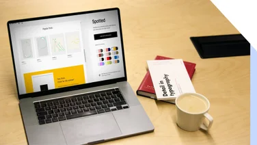 Open laptop displaying a graphic design website with design settings for color palettes, alongside a notebook, tablet, and cup of coffee on a wooden desk.