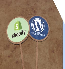 Two signs resting on the sand - one has the Shopify logo, while the other has the WordPress logo