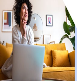 A person in a cozy sweater sits thoughtfully on a yellow couch, focused on a laptop surrounded by a stylish, modern interior.
