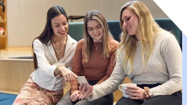 Three women smiling and looking at a smartphone together, with one woman pointing at something on the screen.
