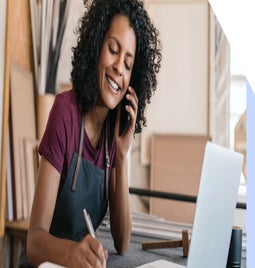 Smiling businesswoman in a t-shirt and apron is on the phone, and makes notes on a pad at her desk, in front of an open laptop