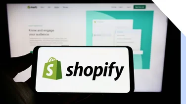 A phone with the shopify logo help in front of a blurred computer screen