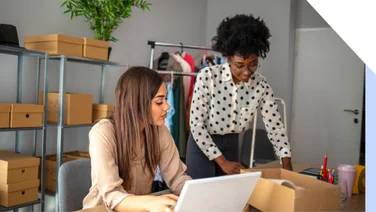 Photo of two women around a laptop and parcel boxes managing their ecommerce site