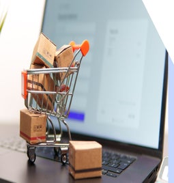 A miniature shopping cart filled with small cardboard boxes rests on a laptop keyboard, with an online shopping interface in the background.