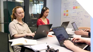 Two women on laptops during a team meeting with sticky notes in the background