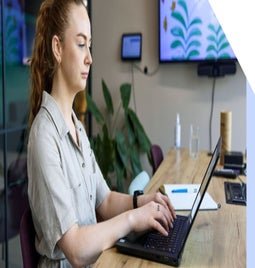 Woman sits in an office working on her laptop with a notebook next to her