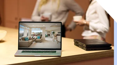 A modern laptop on a wooden table showcasing a virtual house tour with blurred individuals chatting over coffee in the background.