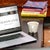 A comfortable reading setup with a laptop open on a blog about books, a cup of coffee, and a stack of books on a wooden table.