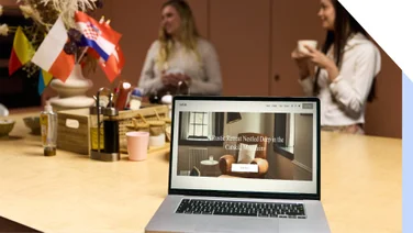 People chat in a kitchen setting with international flags, with a laptop in the foreground showing a rustic retreat in the Catskill Mountains.