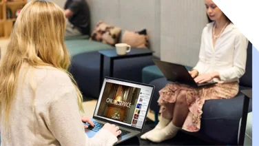 Two women using laptops in a modern office lounge area - one is watching an online service on YouTube.