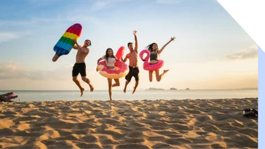 Four people in swimwear jump in the air on a sandy beach holding pool inflatables