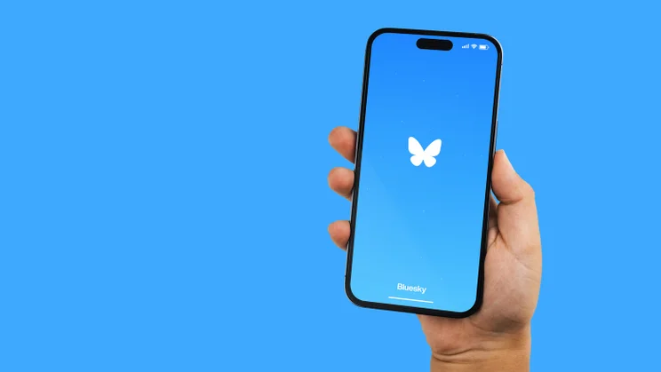 bluesky logo on phone being held in front of a blue background