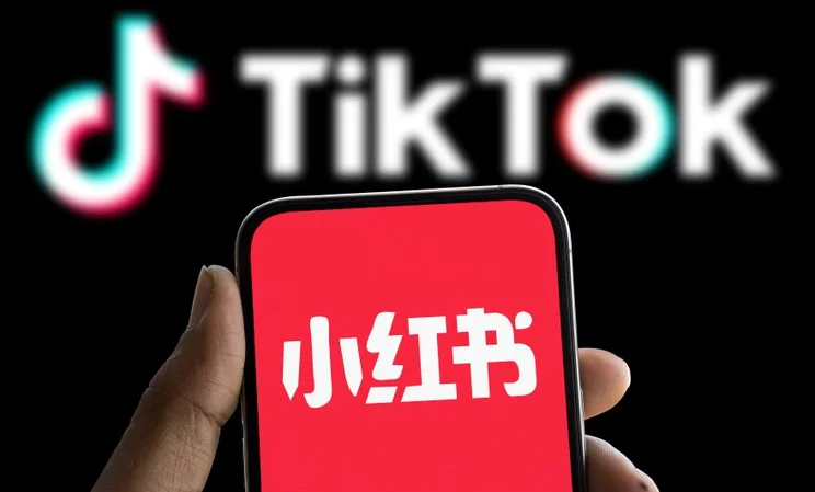 A hand holding a phone with the RedNote logo on screen in front of a monitor with the TikTok logo