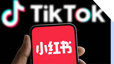 A hand holding a smartphone with the RedNote logo on screen in front of a Monitor showing the TikTok logo