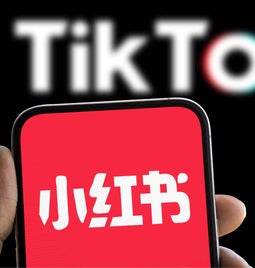 A hand holding a smartphone with the RedNote logo on screen in front of a Monitor showing the TikTok logo