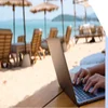 Person working at a laptop on a white sand beach with lounge chairs and umbrellas in the background
