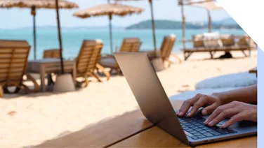 Person working at a laptop on a white sand beach with lounge chairs and umbrellas in the background