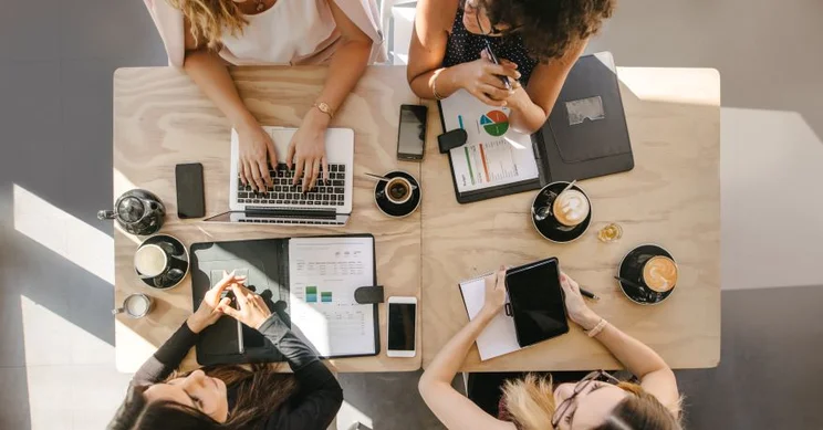Women sat at a desk talking with their work on the table.