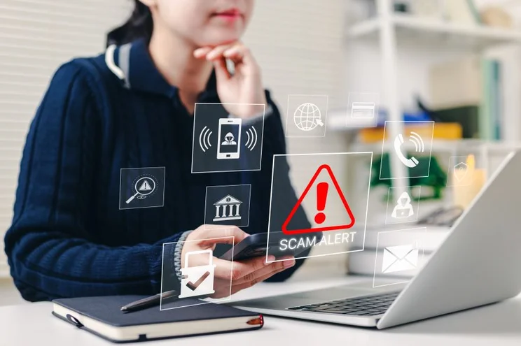 A woman thinking by her laptop with an overlay of a red scam alert symbol