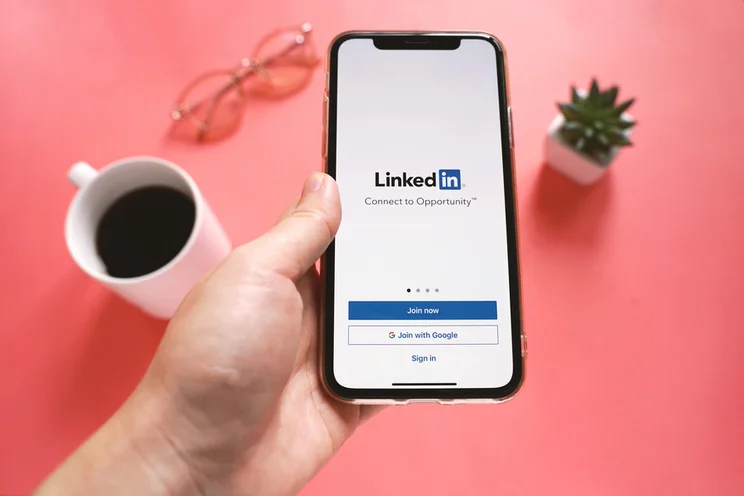 smartphone with linkedin login page on the screen being held over a pink table that has a coffee, a pair of glasses, and a plant on it
