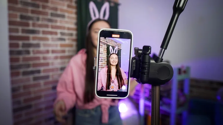 lady in bunny ears and a pink jumper filming herself with a smartphone held up with a tripod