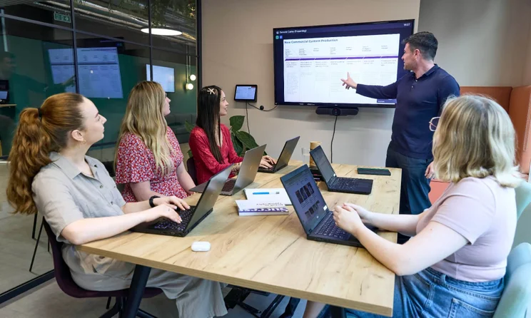 The WBE team sit with laptops around a desk in an office watching a colleague presenting in front of a screen.
