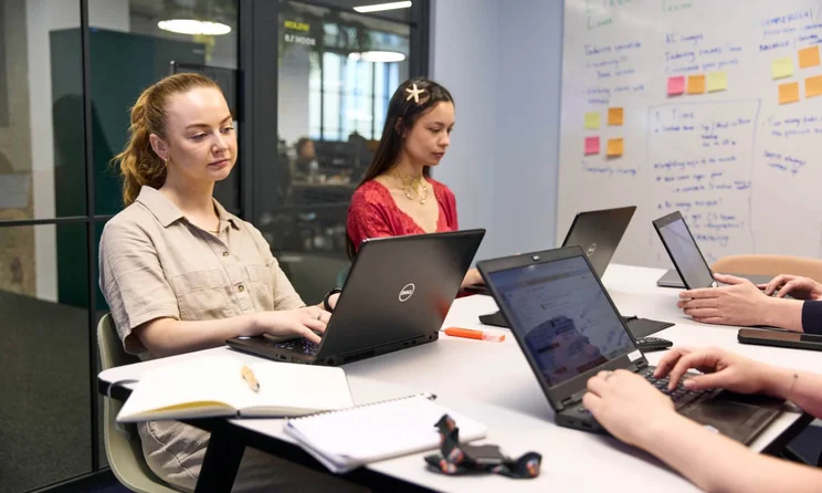 WBE team working on their laptops at a desk in a meeting room; Annie and Holly are visible, concentrating hard