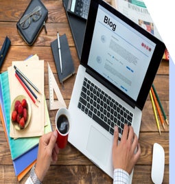 man working on laptop on a wooden table with stationary and paper scattered around him. The laptop is open on a page that says blog