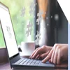 A person types on a laptop in a caf&eacute;, with floating hashtags above, symbolizing social media engagement. A cup is nearby.