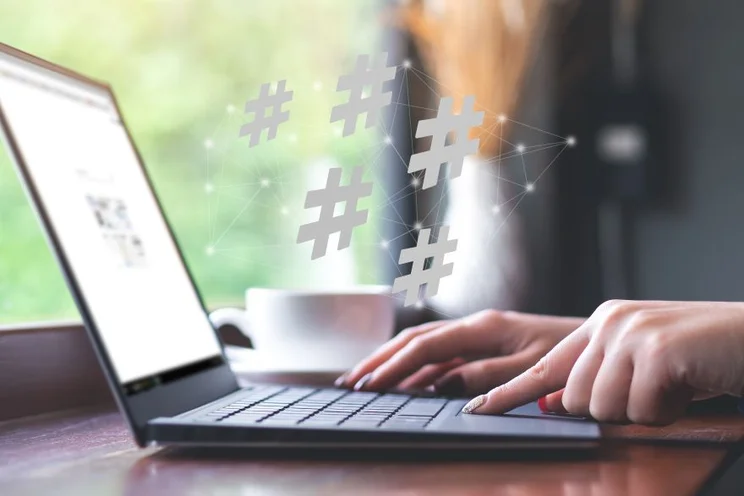 A person types on a laptop in a caf&eacute;, with floating hashtags above, symbolizing social media engagement. A cup is nearby.