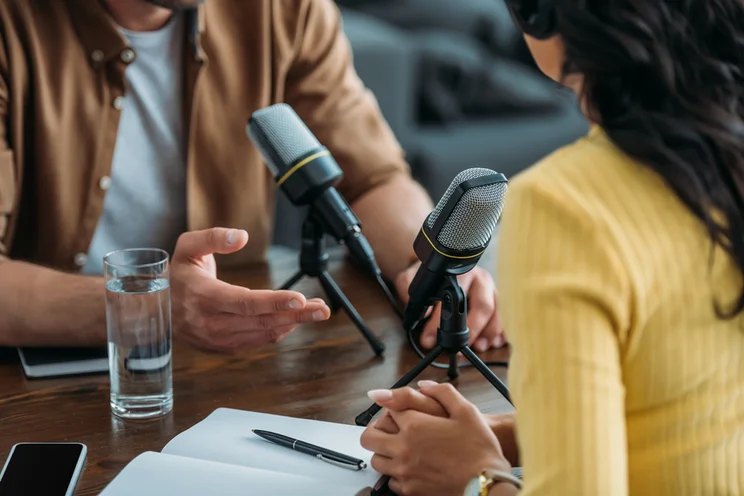 two people sat at wooden table facing each other speaking into microphones