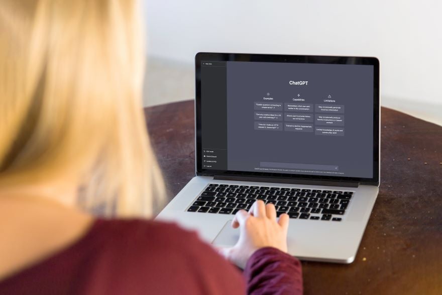 ChatGPT A person with long blonde hair sits at a wooden table, using a laptop displaying the ChatGPT interface with features and limitations.