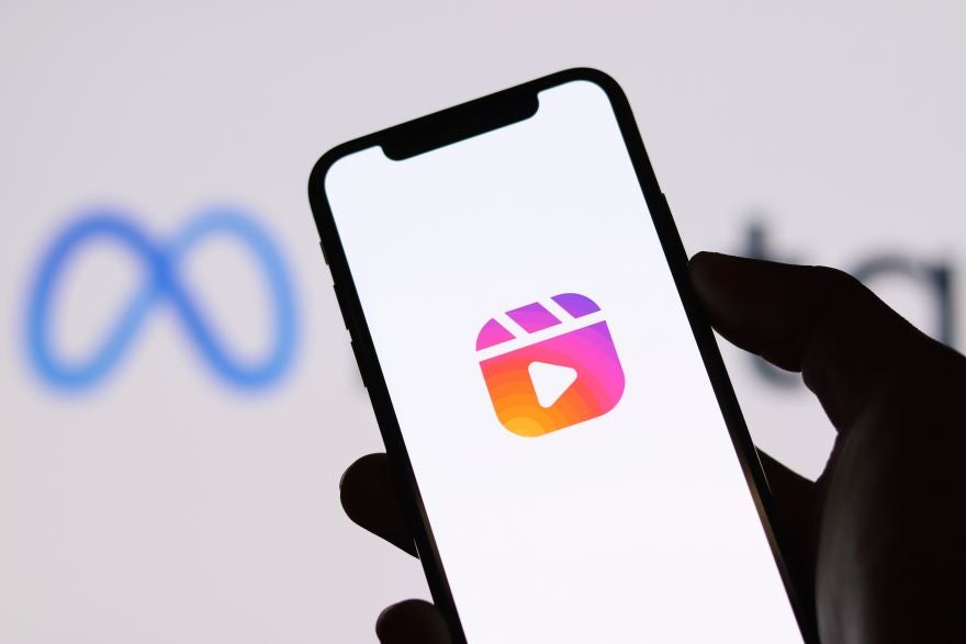 A hand holds a smartphone displaying the Instagram Reels icon with a blurred Meta logo in the background.