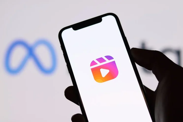 A hand holds a smartphone displaying the Instagram Reels icon with a blurred Meta logo in the background.
