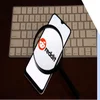 A smartphone displaying the Reddit logo, viewed through a magnifying glass, resting on a keyboard.