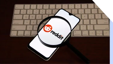 A smartphone displaying the Reddit logo, viewed through a magnifying glass, resting on a keyboard.