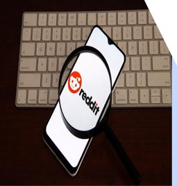 A smartphone displaying the Reddit logo, viewed through a magnifying glass, resting on a keyboard.