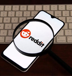 A smartphone displaying the Reddit logo, viewed through a magnifying glass, resting on a keyboard.