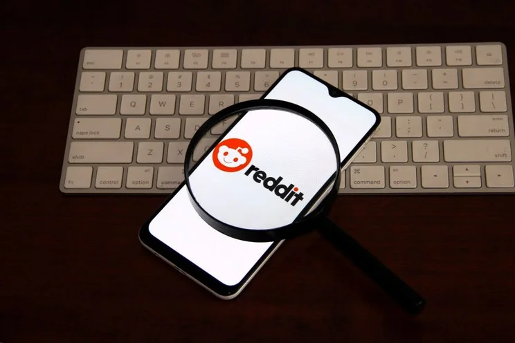A smartphone displaying the Reddit logo, viewed through a magnifying glass, resting on a keyboard.