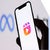A hand holding a smartphone displaying the Instagram Reels logo, with a blurred Meta logo in the background.