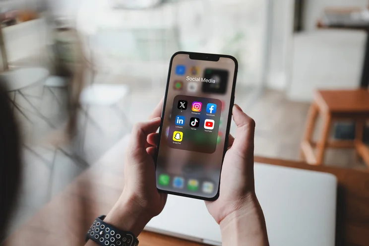 A person holds a smartphone displaying a folder labeled "Social Media" with various app icons, including Instagram, Facebook, TikTok, LinkedIn, Snapchat, and YouTube.