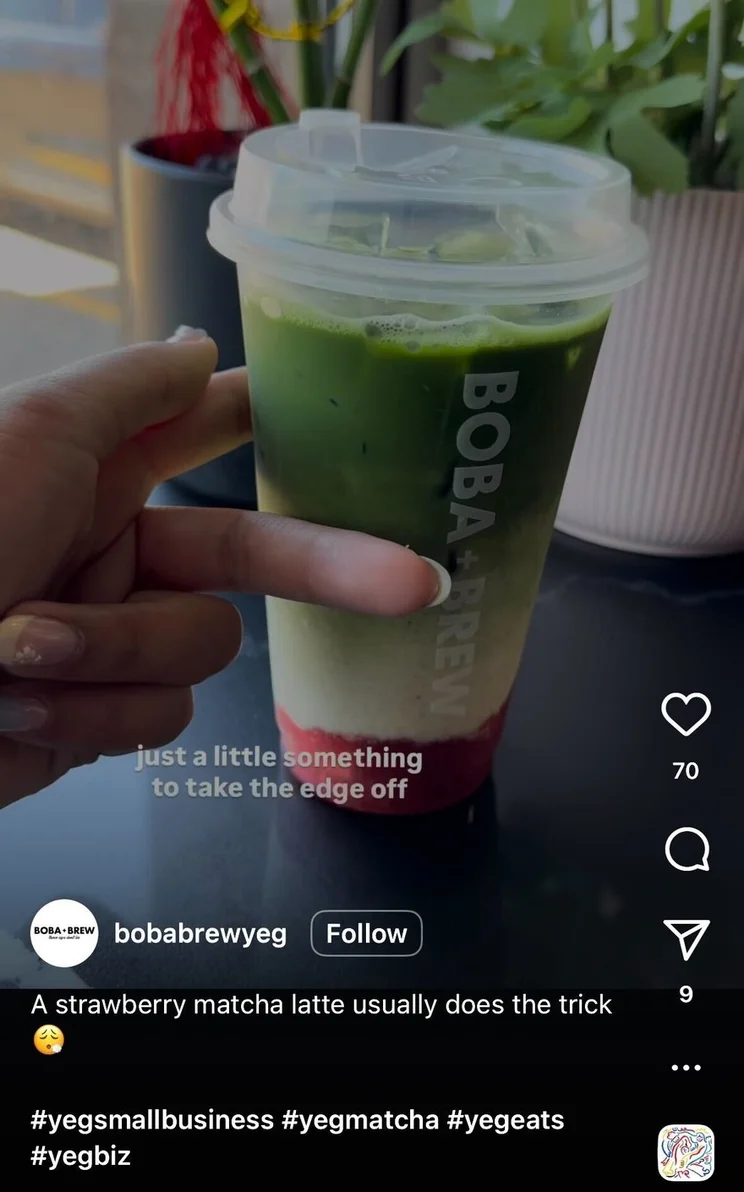 A hand holds a cup of strawberry matcha latte with visible layers of red, white, and green, placed on a dark table next to a leafy plant.
