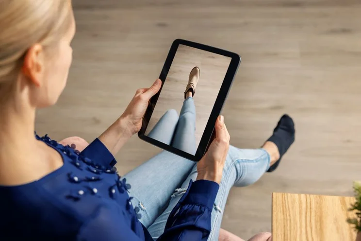 A person holds a tablet displaying a close-up of a shoe worn with jeans, sitting in a cozy, modern setting.