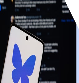 Bluesky logo on a phone, held in front of a screen showing the Bluesky feed