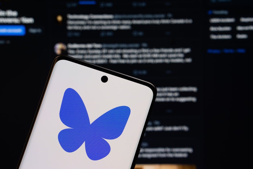 Bluesky logo on a phone, held in front of a screen showing the Bluesky feed