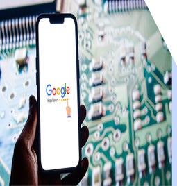 A hand holds a smartphone displaying the Google Reviews logo against a blurred circuit board background.
